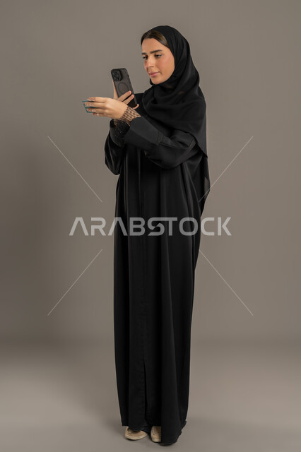 Modern technology and technology, having fun on mobile with friends, browsing social networking sites, portrait of a veiled Emirati Gulf Arab woman wearing an abaya sending text messages on mobile phone, full body, black background