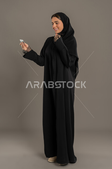 نجاح عملية الشراء والبيع، سيدة عربية خليجية اماراتية محجبة تحمل بيدها بطاقة الإئتمان البنكية، بطاقة الدفع الالكترونية ،  سيده سعوديه تقوم بالدفع الالكتروني عبر كارت المشتريات ،تسديد الفواتير بالبطاقات الإلكترونية، الجسم بالطول الكامل ، خلفية سوداء