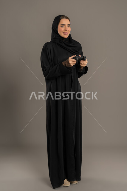 دمج التسلية بالتقنية والتكنولوجيا، شابة سعودية مبتسمة تلعب بايماءات الاندماج والتفاعل، قضاء اوقات ممتعة  بألعاب الفيديوز الترفيهية في عطلة نهاية الاسبوع ، بورتريه لإمرأة عربية خليجية اماراتية ترتدي العباءة تحمل بيدها عصا التحكم، الجسم بالطول الكامل, خلفية سوداء