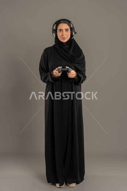 تجارب ترفيهية ممتعة ومسلية, ايماءات الاندماج والتفاعل أثناء اللعب, قضاء إجازة صيفية ممتعة, بورتريه لإمرأة عربية خليجية إماراتية محجبة ترتدي العباءة تضع سماعة الهيدفون وتحمل بيدها عصا التحكم، شابة سعودية تلعب بألعاب الفيديو الترفيهية، الطول الكامل للجسم، خلفية سوداء