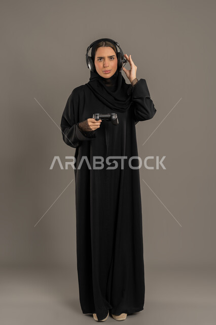 إيماءات الاستمتاع والاندماج والتفاعل, قضاء إجازة صيفية ممتعة، تقنيات الكترونية حديثة, بورتريه لإمرأة عربية خليجية إماراتية محجبة ترتدي العباءة تضع سماعة الهيدفون وتحمل بيدها عصا التحكم، شابة سعودية تلعب بألعاب الفيديو الترفيهية، الطول الكامل للجسم، خلفية بيضاء