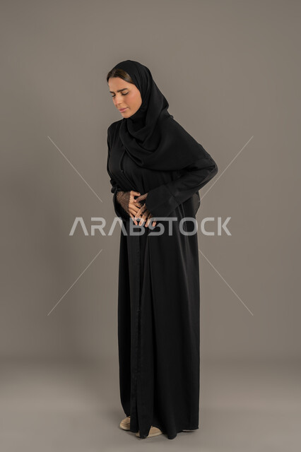 حركات وجه ويدين تدل على الاضطراب والانزعاج، ضرورة الراحة وأخذ العلاج المناسب، بورتريه لامراة عربية خليجيه سعودية محجبة ترتدي العباءة السوداء تقوم بوضع يديها على بطنها بتعابير التألم, سيدة سعوديه تشعر بالمرض ووجع في البطن، الجسم بالطول الكامل، خلفية سوداء