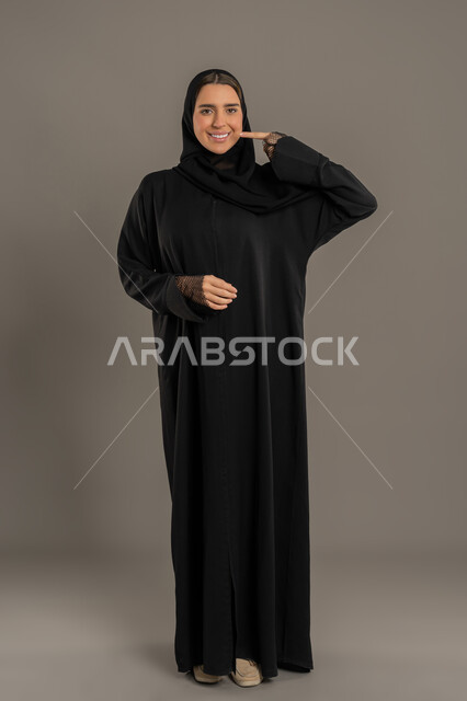 Illustration and indication of a specific place, pointing to an item, portrait of a veiled Emirati Gulf Arab woman wearing a black abaya pointing with her index finger, smiling young Saudi woman locating something, full body, black background