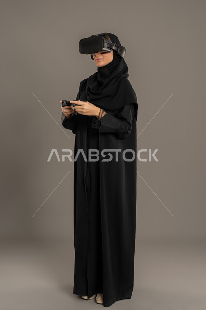 المتعة و التسلية بألعاب الفيديو والعالم الافتراضي، استخدام جهاز تقني حديث ومتطور، بورتريه لإمراة عربية خليجية اماراتية محجبة ترتدي العباءة السوداء ونظارات ثلاثية الأبعاد، سيده سعودية تقف وتحمل بيدها عصا التحكم، تعابير وايماءات الاندماج، الجسم بالطول الكامل، خلفية سوداء