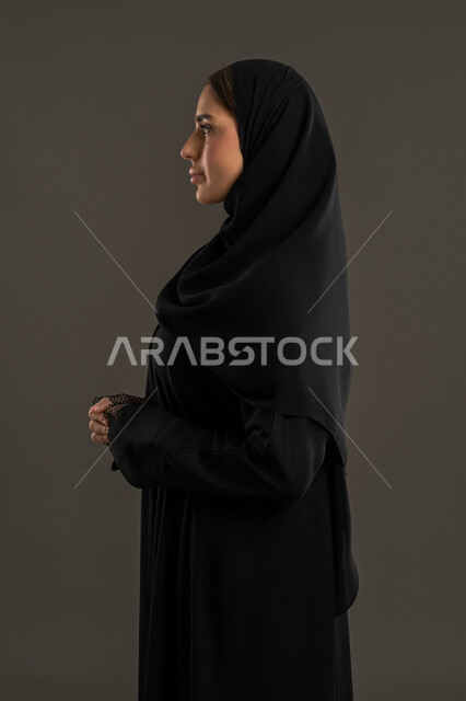 الاهتمام بالمظهر الخارجي والاناقة, ايماءات الثقة بالنفس ، الاناقة والاهتمام بالمظهر الخارجي, بورتريه من الجانب لامراة عربية خليجية إماراتية ترتدي العباءة السوداء والحجاب تقف باستقامة ، سيدة سعودية محجبة تنظر الى شئ ما بتعابير السعادة, خلفية سوداء