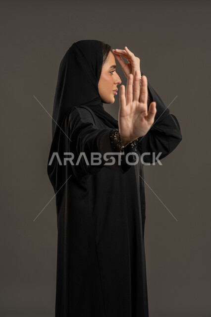 الامتناع عن فعل أمر معينا , بورتريه من الجانب لسيدة عربية خليجية امارتيه محجبه ترتدي العباءه السوداء  تغطي وجهها بيدها و تشير بكفة يدها الأخرى بإيماءات و تعابير وجه و يدين تدل على التوقف و الابعاد،شابة سعودية تشعر بالإنزعاج و الرفض والإمتناع, خلفية سوداء