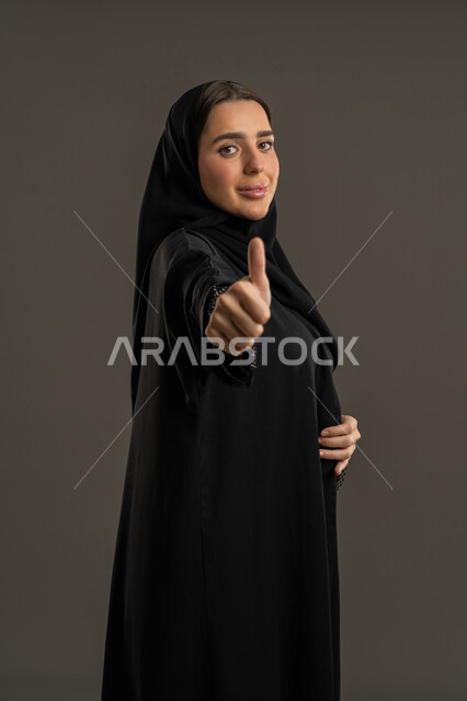 علامة الامتياز و الجودة، النظر الى الكاميرا بتعابير السرور, سيدة سعودية مبتسمه تشعر بالإعجاب والرضى، بورتريه لامراة عربية خليجية اماراتية مبتسمة محجبة ترتدي العباءة السوداء ترفع اصبع الابهام للأعلى بإيماءات القبول و الموافقة، خلفية سوداء