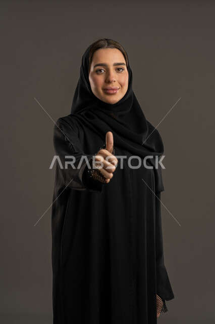 مفهوم القبول والموافقه، النظر الى الكاميرا بتعابير السرور, سيدة سعودية مبتسمه تشعر بالإعجاب والرضى، بورتريه لامراة عربية خليجية اماراتية مبتسمة محجبة ترتدي العباءة السوداء ترفع اصبع الابهام للأعلى بإيماءات القبول و الموافقة، خلفية سوداء