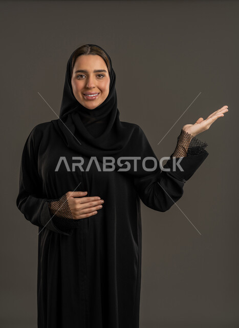 Hospitality and warm welcome, looking at the camera with expressions of pleasure and cheerfulness, gestures of peace and welcome, close-up portrait of a veiled Emirati Gulf Arab woman wearing a black abaya and raising her hand up, a Saudi woman welcoming someone, black background