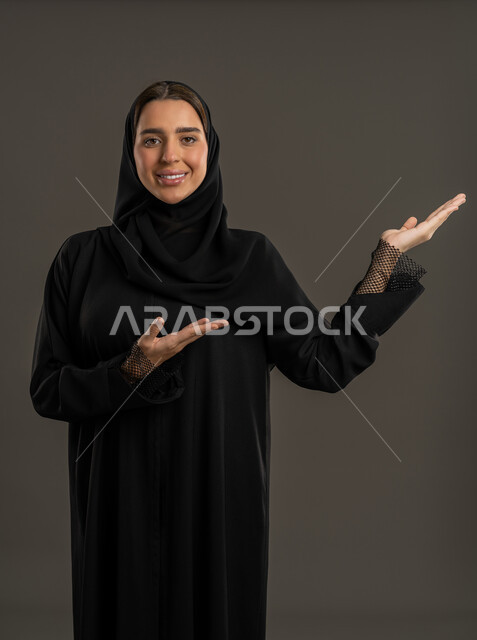 Welcoming and receiving cheerfully, looking at the camera with expressions of pleasure and cheerfulness, gestures of peace and welcome, close-up portrait of a veiled Emirati Gulf Arab woman wearing a black abaya and raising her hand up, a Saudi woman welcoming someone, black background