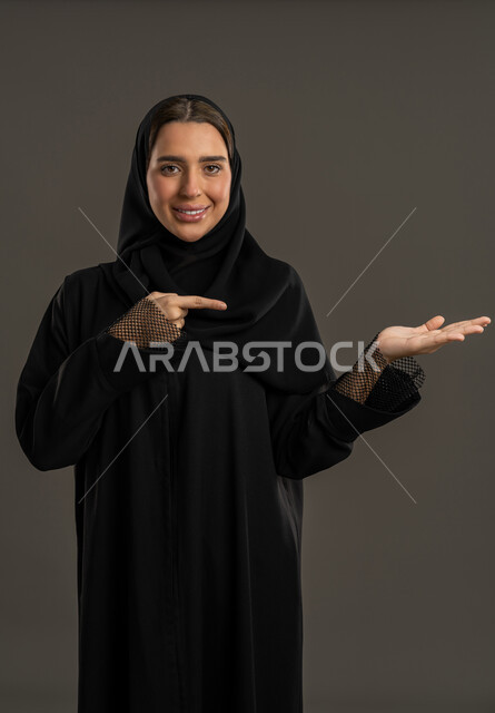 إشارات وحركات يدين تدل على هذا المكان (هنا)، صورة مقربة لامرأة عربيه خليجيه اماراتية مبتسمه محجبه ترفع اصابعها السبابة وتشير الى شيء ما، سيده سعوديه تقوم بتحديد موقع معين بإيماءات البشاشة، خلفية سوداء