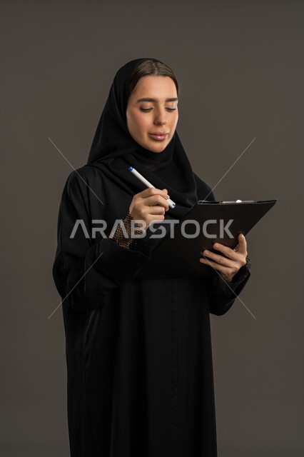 تدوين و كتابة تقارير أبحاث علمية، سيده سعودية تقوم بتدوين الملاحظات وإعداد التقرير اليومي، بورتريه لامراة عربية خليجيه اماراتية تمسك بيدها سنادة ورق سوداء وقلم تنظر اليها بإيماءات التركيز والتمعن، خلفيه سوداء
