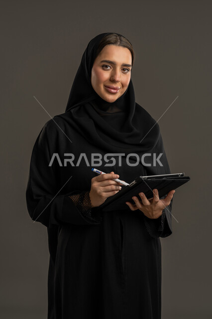 مفهوم إدارة المشاريع عن بعد، متابعة المراسلات والسجلات، بورتريه لامراة عربية خليجية اماراتية محجبة ترتدي العباءة السوداء تحمل بيديها حامل اوراق تقوم بتدووين الملاحظات ، سيده سعودية مبتسمة تقوم بانجاز المهام المطلوبة، خلفية سوداء