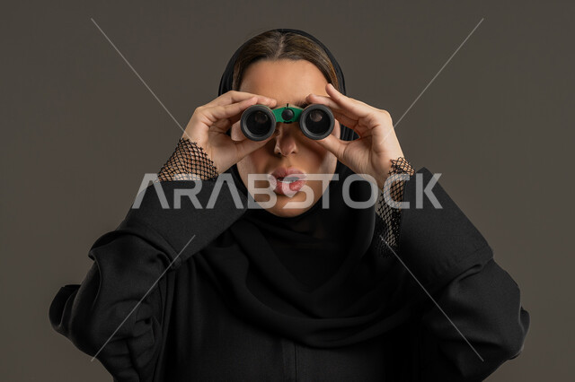 الرؤية بالعدسة المكبرة، حب الاستكشاف والمغامرة، بورتريه مقرب لامرأة عربية خليجية اماراتية محجبة ترتدي العباءة السوداء تنظر من خلال منظار بايماءات الدهشه و الاستغراب, شابة سعودية  تقوم برؤية الاشياء البعيدة عبر التلسكوب، خلفية سوداء