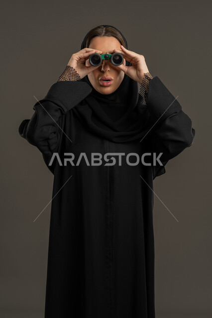 التخطيط المستقبلي للأهداف و الطموحات، حب الاستكشاف والمغامرة، بورتريه لامرأة عربية خليجية اماراتية محجبة ترتدي العباءة السوداء تنظر من خلال منظار بايماءات الدهشه و الاستغراب, شابة سعودية  تقوم برؤية الاشياء البعيدة عبر التلسكوب، خلفية سوداء