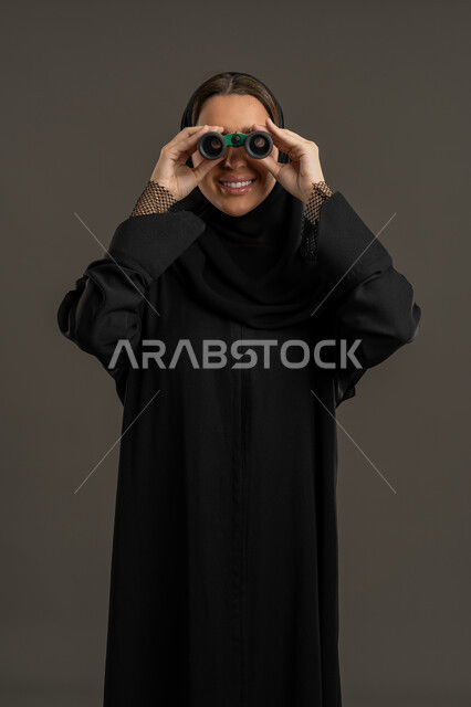 حب المغامرة و الاستكشاف، رؤية الاشياء البعيدة عبر التلسكوب، سيده سعوديه تقوم بالتخطيط المستقبلي للأهداف و الطموحات، بورتريه لامرأة عربية خليجية اماراتية مبتسمة محجبة ترتدي العباءة السوداء تنظر من خلال منظار ثنائي العينين, خلفية سوداء