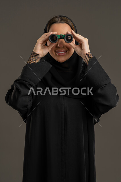 مفهوم الاستكشاف والمغامرة، رؤية الاشياء البعيدة عبر التلسكوب، التخطيط المستقبلي للأهداف و الطموحات، بورتريه لامرأة عربية خليجية اماراتية مبتسمة محجبة ترتدي العباءة السوداء تنظر من خلال منظار ثنائي العينين, خلفية سوداء