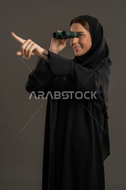 التخطيط المستقبلي للأهداف والطموحات، التفكير والتطلع إلى المستقبل، رؤية الأشياء البعيدة، حب الاستكشاف والمغامرة، بورتريه لشابة عربية خليجية اماراتية محجبة ترتدي العباءة تمسك منظار ثنائي العينين، امراة سعودية تقوم بالاشارة باصبع السبابة الى مكان ما ,خلفية سوداء