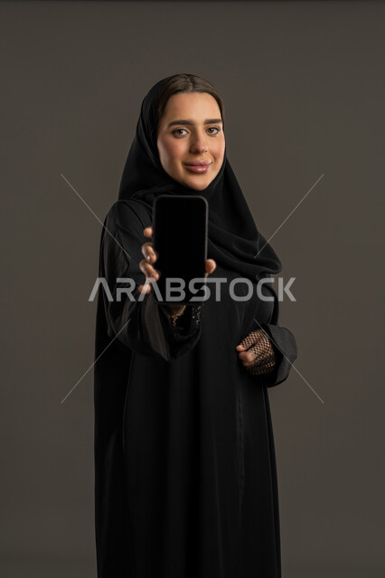 عرض خدمات وتطبيقات الجوال , استخدام الأجهزة التقنية الحديثة والمتطورة، بورتريه لامرأة عربية خليجية اماراتية مبتسمة ترتدي الحجاب والعباءة تعوم بعرض شاشة سوداء فارغة,  سيده سعودية تحمل بيدها الهاتف المحمول تنظر إلى الكاميرا بإيماءات الثقة بالنفس، خلفية سوداء