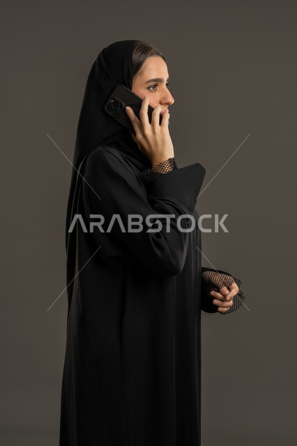 Expressions of immersion and concentration while talking on the mobile phone, having a good time exchanging news and conversations, side portrait of a veiled Emirati Gulf Arab woman wearing a black abaya holding a mobile phone in her hand, smiling young Saudi woman making a phone call, black background