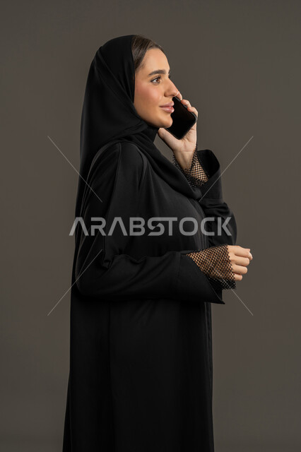قضاء اوقات ممتعة بتبادل الاخبار والأحاديث, تفاعل اجتماعي عبر الأجهزة الذكية, بورتريه من الجانب لامرأة عربية خليجية إماراتية محجبة ترتدي العباءة السوداء تمسك بيدها الهاتف المحمول, شابة سعودية مبتسمة تقوم بإجراء اتصال هاتفي, خلفية سوداء