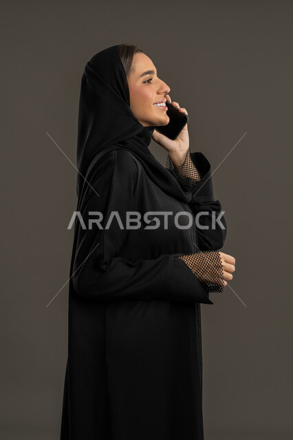 مشاركة الأخبار والأحاديث الجيدة, التواصل مع العائلة والأصدقاء, استخدام الأجهزة التقنية الحديثة في الحياة اليومية, بورتريه من الجانب لامرأة عربية خليجية إماراتية محجبة ترتدي العباءة السوداء تمسك بيدها الهاتف المحمول, شابة سعودية مبتسمة تقوم بإجراء اتصال هاتفي, خلفية سوداء