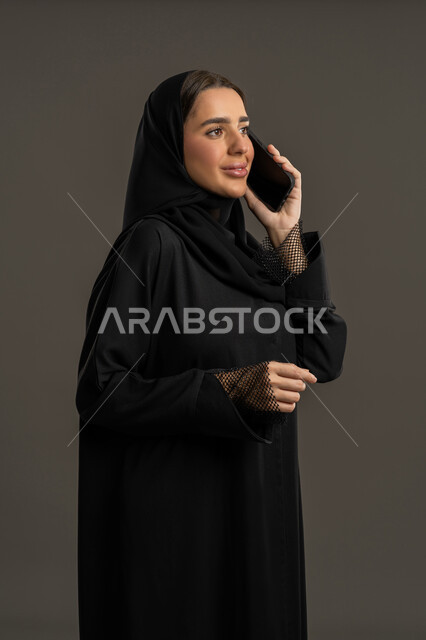 استخدام الأجهزة التقنية الحديثة في الحياة اليومية,التواصل مع العائلة والأصدقاء, مشاركة الأخبار والأحاديث الجيدة, بورتريه لامرأة عربية خليجية إماراتية محجبة ترتدي العباءة السوداء تمسك بيدها الهاتف المحمول, شابة سعودية تقوم بإجراء اتصال هاتفي, خلفية سوداء