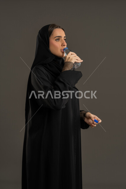 المحافظة على سلامة الجهاز المناعي للجسم ,بورترية لامراة عربية خليجية امارتية محجبة ترتدي العباءة السوداء تقوم بالشرب من زجاجة ماء بلاستيكية صغيرة, سيدة سعودية تكثر من السوائل للحفاظ على الجسد من الجفاف , خلفية سوداء