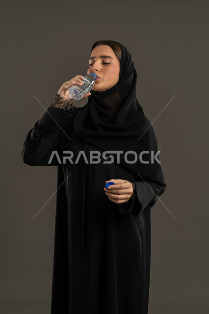 Drinking water regularly, maintaining the health of the body's immune system, portrait of a veiled Emirati Gulf Arab woman wearing a black abaya drinking from a small plastic water bottle, a Saudi woman feeling thirsty and dehydrated, black background