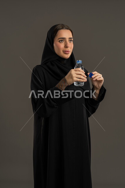 التخلص من سموم الجسم، أهمية شرب الماء النقي للمحافظة على بشرة نضرة, بورتريه لإمرأة عربية خليجية اماراتية ترتدي الحجاب و العباءة السوداء تحمل بيدها زجاجة ماء, سيدة سعودية تقوم بفتح زجاجه بلاستيكية, الشعور بالعطش والجفاف, خلفية سوداء