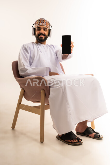 Using modern technology and techniques, entertainment and recreation in free time, portrait of a young Arab Gulf Omani man wearing a dishdasha and a kuma, holding a mobile phone in his hand, showing a blank black screen, full-length body, white background.