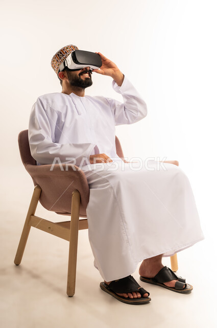 استخدام التقنية والاجهزة الحديثة ، ارتداء نظارة الفي آر ثلاثية الأبعاد, بورتريه لشاب عربي خليجي عماني يرتدي الدشداشة والكمة يجلس على كرسي مريح ينظر من خلال نظارات الواقع الافتراضى, رفع كف اليد للأعلى بإيماءات الاندماج ومحاكاة العالم الخيالي، خلفية بيضاء .