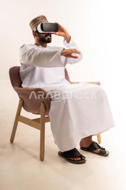 Wearing 3D VR glasses, technical and technological development, portrait of a young Arab Gulf Omani man wearing a dishdasha and a kuma sitting on a comfortable chair looking through virtual reality glasses, raising his palm upwards in a gesture of integration and simulating the imaginary world, white background.