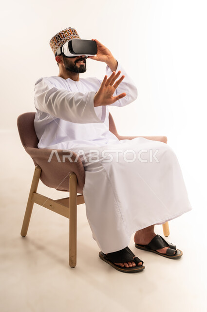 التطور التقني والتكنولوجي, المتعة بالمشاهدة عبر نظارة الواقع الافتراضي، بورتريه لشاب عربي خليجي عماني يرتدي الدشداشة والكمة يجلس على كرسي مريح ينظر من خلال نظارات ثلاثية الأبعاد (الفي ار), رفع كف اليد للأعلى بإيماءات الاندماج ومحاكاة العالم الخيالي، خلفية بيضاء .