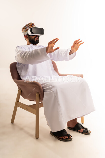 المتعة بالمشاهدة عبر نظارة الواقع الافتراضي، التطور التقني والتكنولوجي, بورتريه لشاب عربي خليجي عماني يرتدي الدشداشة والكمة يجلس على كرسي مريح ينظر من خلال نظارات ثلاثية الأبعاد (الفي ار), رفع كف اليد للأعلى بإيماءات الاندماج ومحاكاة العالم الخيالي، خلفية بيضاء .