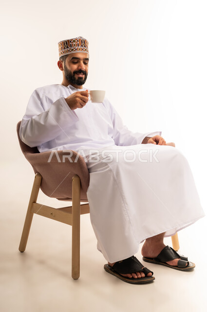 تناول الأعشاب الشتوية الدافئة، قضاء وقت الفراغ باحتساء المشروب المفضل, الجلوس والاستمتاع بالمشروبات الساخنة , بورتريه لشاب عربي خليجي عماني مبتسم يرتدي الكمة و الدشداشة يجلس على كرسي مريح يقوم بشرب القهوة بإيماءات السعادة و الفرح, خلفية بيضاء .