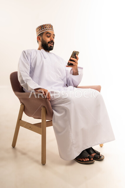 الدردشة مع الاهل والأصدقاء عبر تطبيقات الجوال، استقبال خبر مفاجئ ،  تصفح مواقع التواصل الاجتماعي, بورتريه لرجل عربي خليجي عماني يرتدي الدشداشة والعمامة يجلس على كرسي مريح يحمل بيده الهاتف المحمول, استخدام التقنيات الحديثة المتطورة, خلفية بيضاء .
