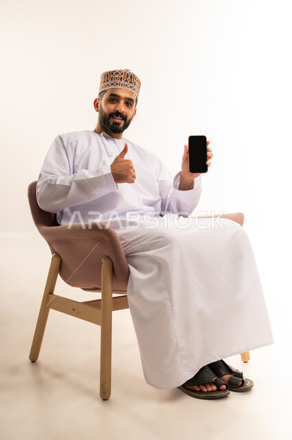 Using advanced modern technology and techniques, portrait of a smiling young Omani Arab Gulf man wearing a dishdasha and a kummah sitting on a comfortable chair, displaying a blank black screen via mobile phone, looking at the camera with expressions of pleasure and quality, full body image, white background.