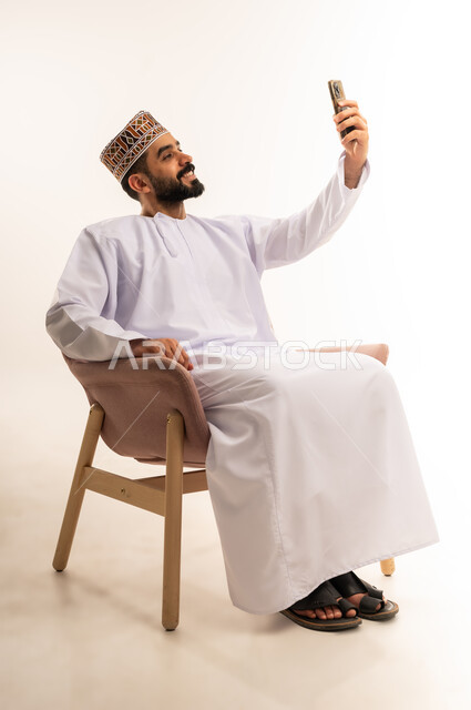 توثيق اللحظات السعيدة عبر الهاتف المحمول, استخدام جهاز تقني حديث ومتطور, بورتريه لشاب عربي خليجي عماني يرتدي الدشداشة والكمة يجلس على كرسى مريح ، يقوم بالتقاط صورة السيلفي التذكارية عبر الجوال, خلفية بيضاء .