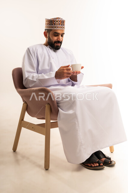 Sitting and enjoying hot drinks and warm winter herbs, spending free time sipping your favorite drink, portrait of a smiling young Arab Gulf Omani man wearing a keffiyeh and a dishdasha sitting on a comfortable chair drinking coffee with gestures of happiness and joy, white background.