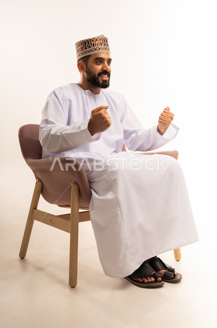 ايماءات الفوز والانتصار ، قضاء وقت ممتع بمتابعة المباراة، بورتريه لرجل عربي خليجي عماني يرتدي الدشداشة والكمة يجلس على أريكة مريحة معه كرة القدم يرفع قبضة يده لاعلى بتعابير الحماس ، المباريات والألعاب الرياضية، خلفية بيضاء