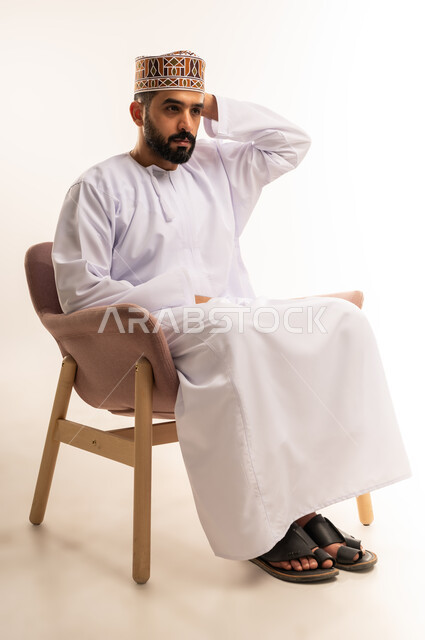 مشاهدة ومتابعة مباراة كرة القدم، ايماءات الحزن والخسارة ،بورتريه لرجل عربي خليجي عماني يرتدي الدشداشة والكمة يجلس على كرسى مريح  يضع يديه خلف رأسه بايماءات الهزيمة ، المباريات والألعاب الرياضية، خلفية بيضاء .