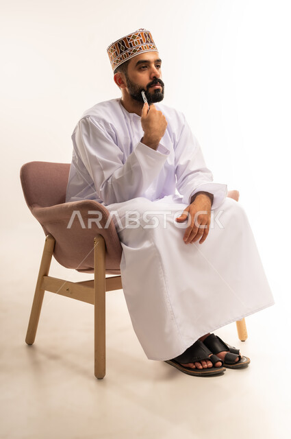 Planning to solve a problem, feeling confused and uncertain. A portrait of an Omani Arabian Gulf man wearing a dishdasha and a kummah sitting on a chair, his hand under his chin, looking at something with an expression of thought. Gestures of contemplation, reflection, and concentration. Full-length body, against a white background.