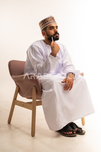 إيماءات التأمل والتمعن والتركيز, بورتريه لرجل عربي خليجي عماني يرتدي الدشداشة والكمة يجلس على كرسى ويضع يده تحت ذقنه، النظر الى مكان ما بتعابير التفكير، التخطيط لحل مشكلة ما، الشعور بالحيرة وعدم التأكد, الجسم بالطول الكامل، خلفية بيضاء .