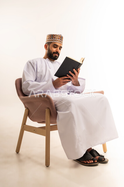 ممارسة هواية المطالعة, التعلم والتثقيف من خلال قراءه الكتب و الروايات  الهادفه، بورتريه لشاب عربي خليجي عماني يرتدي الكمة و الدشداشة يجلس على كرسي مريح يمسك بيده كتاب بإيماءات التمعن والتركيز, خلفيه بيضاء .