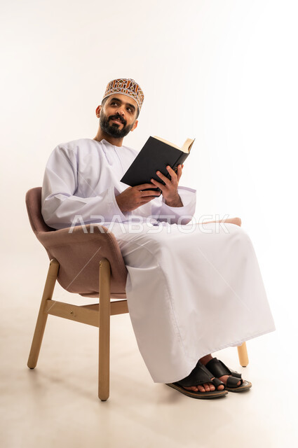 التعلم والتثقيف من خلال قراءه الكتب و الروايات  الهادفه، ممارسة هواية المطالعة, بورتريه لشاب عربي خليجي عماني يرتدي الكمة و الدشداشة يجلس على كرسي مريح يمسك بيده كتاب وينظر الى شئ ما بإيماءات التفكير و الحيرة, خلفيه بيضاء .