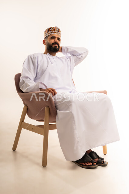 الإحساس بالإرهاق و الكسل والخمول, بورتريه لرجل عربي عماني خليجي يرتدي الدشداشة والكمة يجلس على كرسي, إيماءات وجه ويدين بالحاجه الى الراحه , الشعور بالتعب والنعاس و النوم, خلفية بيضاء .