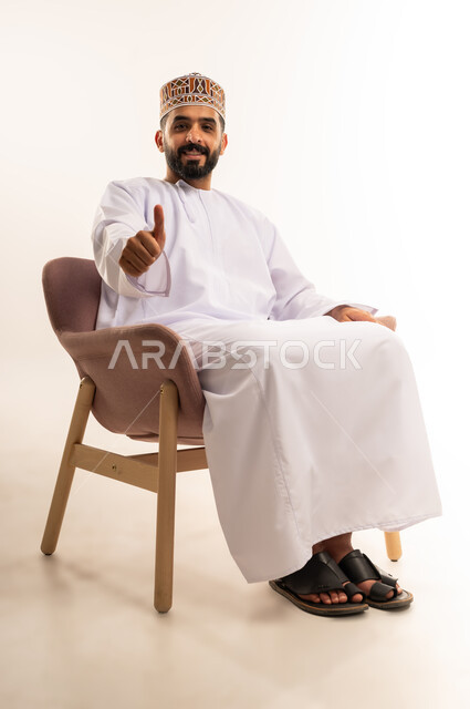 تعابير الموافقة والرضى والقبول، بورتريه لشاب عربي خليجي عماني مبتسم يرتدي الدشداشة والكمة يرفع اصبع الابهام للاعلى بإيماءات الاعجاب والسرور،علامة الجودة و الامتياز، خلفية بيضاء .