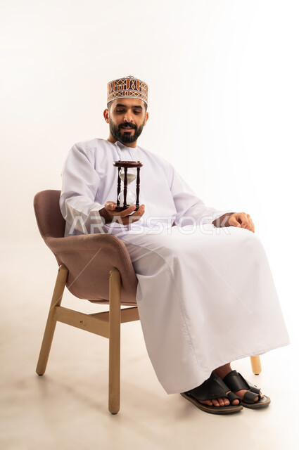 مفهوم إدارة الأعمال وتنظيم الجدول الزمني, إدراك قيمة اللحظات الثمينة, القيام بمراقبة توقيت الرمل, بورتريه لشاب عربي خليجي عماني يرتدي الكمة و الدشداشة يجلس على كرسي مريح يمسك بيده الساعة الرملية الزجاجية (البنكام) وينظر إليها, خلفية بيضاء .