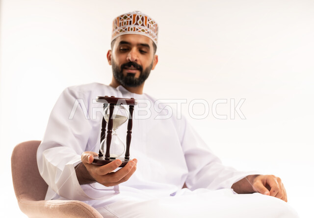 القيام بمراقبة توقيت الرمل, مفهوم إدارة الأعمال وتنظيم الجدول الزمني, إدراك قيمة اللحظات الثمينة, بورتريه مقرب لشاب عربي خليجي عماني يرتدي الكمة و الدشداشة يجلس على كرسي مريح يمسك بيده الساعة الرملية الزجاجية (البنكام) وينظر إليها, خلفية بيضاء .